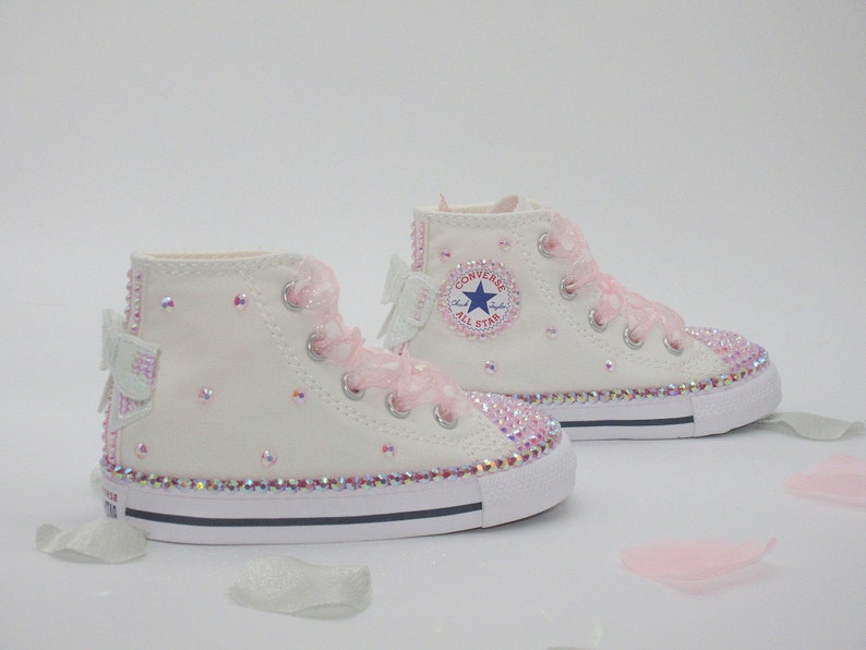 May include: White high-top Converse sneakers with pink ribbon bows and rhinestones. The sneakers have a blue star logo and the text "Converse All Star" on the side.