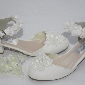 May include: White girls' dress shoes with a low heel and a white flower embellishment on the ankle strap. The shoes have a sparkly white finish and a white lace flower design on the toe.