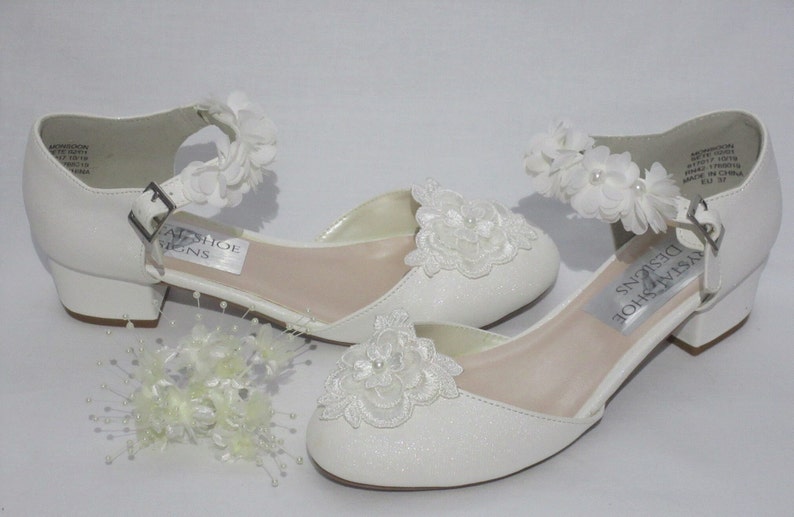 May include: White girls' dress shoes with a low heel and a white flower embellishment on the ankle strap. The shoes have a sparkly white finish and a white lace flower design on the toe.