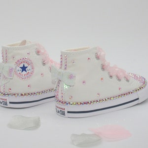 May include: White high-top Converse sneakers with pink bows and rhinestones. The shoes have a white canvas upper, a black rubber sole, and a white rubber toe cap. The Converse logo is visible on the side of the shoe.