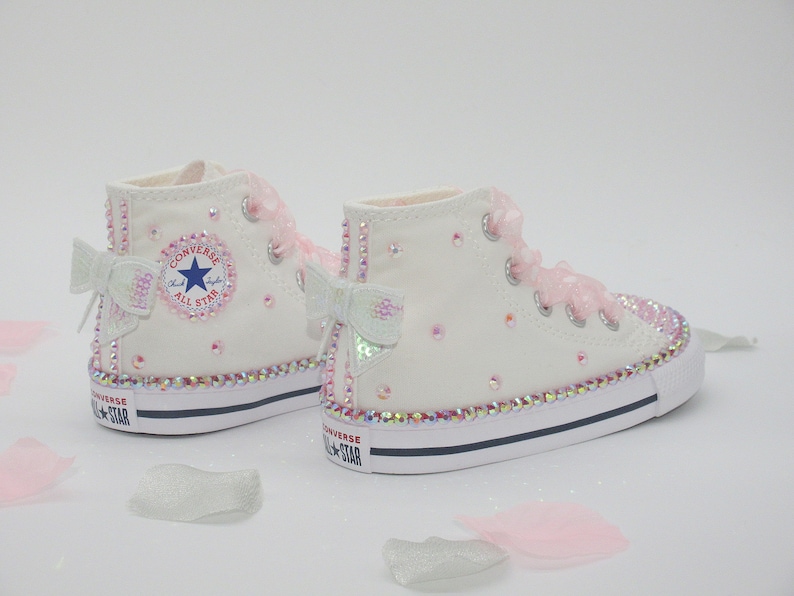 May include: White high-top Converse sneakers with pink bows and rhinestones. The shoes have a white canvas upper, a black rubber sole, and a white rubber toe cap. The Converse logo is visible on the side of the shoe.