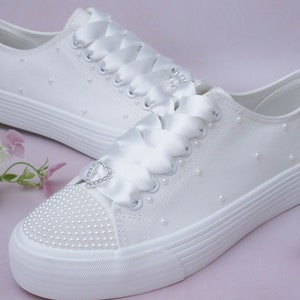 Può includere: Sneakers bianche in tela con un nastro di raso legato intorno alla caviglia. Le scarpe sono decorate con perle e un piccolo cuore di strass.