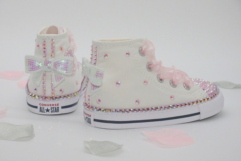 May include: White high-top Converse sneakers with pink and iridescent bows and rhinestones. The shoes have the text "Converse All Star" on the back.