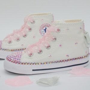 May include: White high-top Converse sneakers with pink ribbon laces and a sparkly pink trim. The shoes are decorated with iridescent rhinestones.