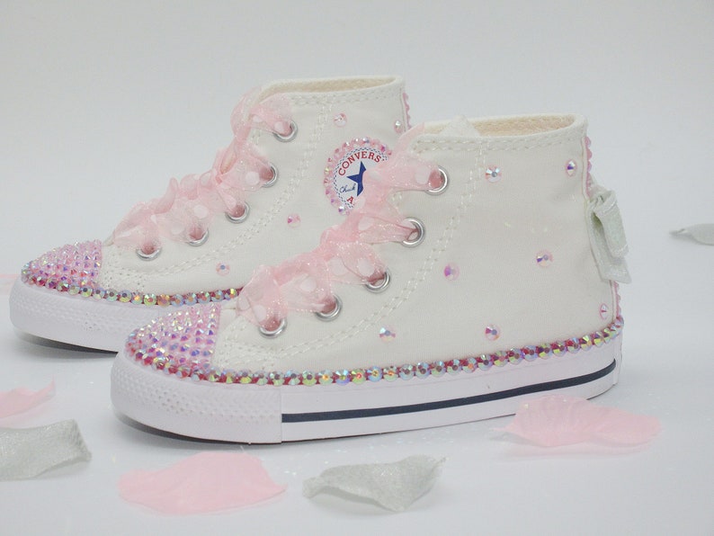 May include: White high-top Converse sneakers with pink ribbon laces and a sparkly pink trim. The shoes are decorated with iridescent rhinestones.