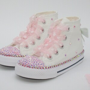 May include: White high-top sneakers with pink polka dot ribbon laces and a sparkly pink trim. The shoes are decorated with iridescent rhinestones.