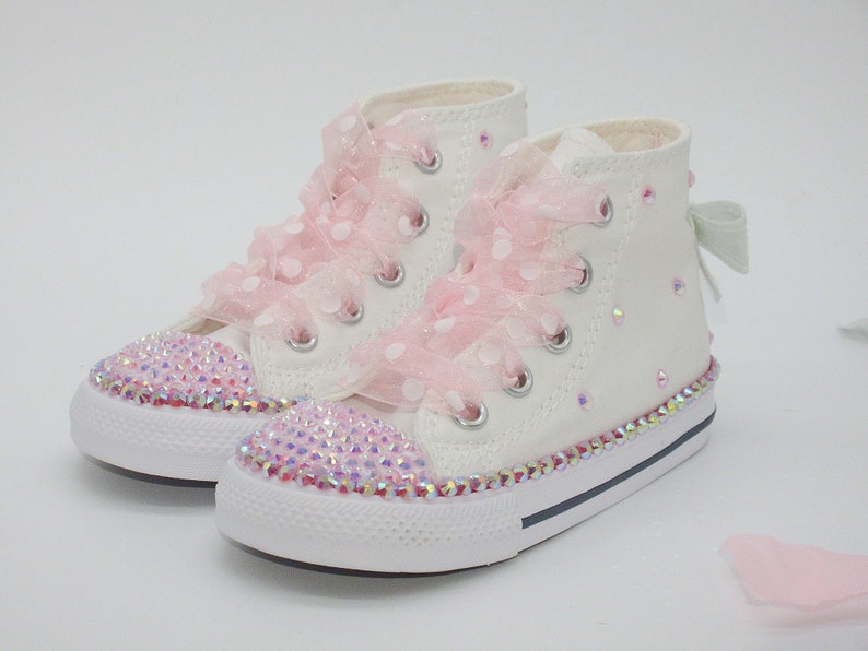 May include: White high-top sneakers with pink polka dot ribbon laces and a sparkly pink trim. The shoes are decorated with iridescent rhinestones.