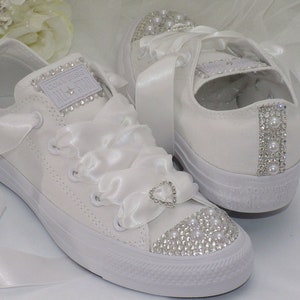 May include: White Converse All-Star trainers decorated with pearls and rhinestones. The toe caps are covered in small rhinestones and pearls. White satin ribbons replace the laces. A heart-shaped rhinestone embellishes the front.