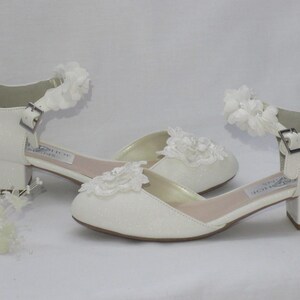 May include: White bridal shoes with a low block heel and a delicate lace flower embellishment on the toe and ankle strap.
