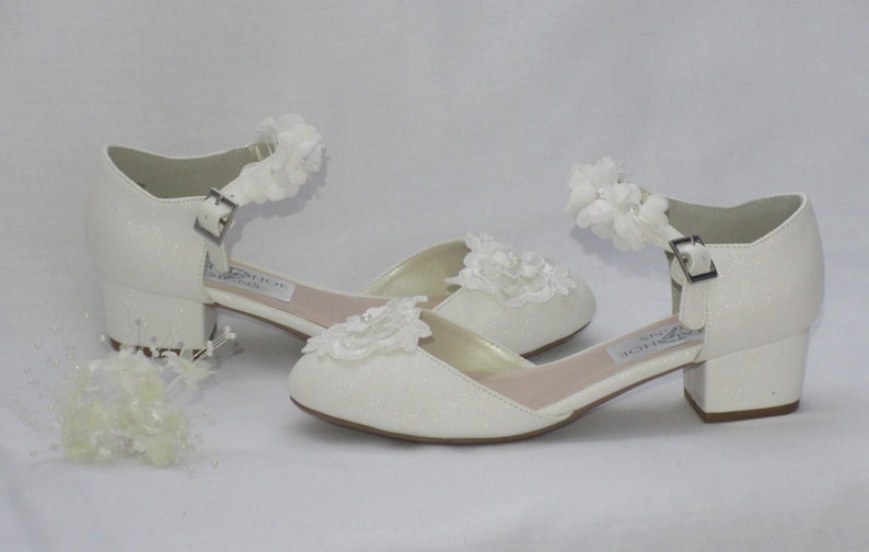 May include: White bridal shoes with a low block heel and a delicate lace flower embellishment on the toe and ankle strap.