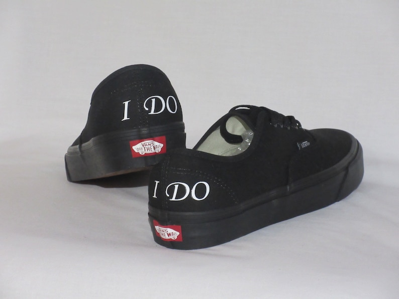 May include: Black Vans trainers with the words "I DO" in white on the heels. The shoes have black soles and the Vans logo on the back. A classic, casual style of footwear.