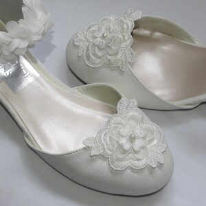 May include: White satin bridal shoes with delicate lace flower embellishments and an ankle strap adorned with white fabric flowers.