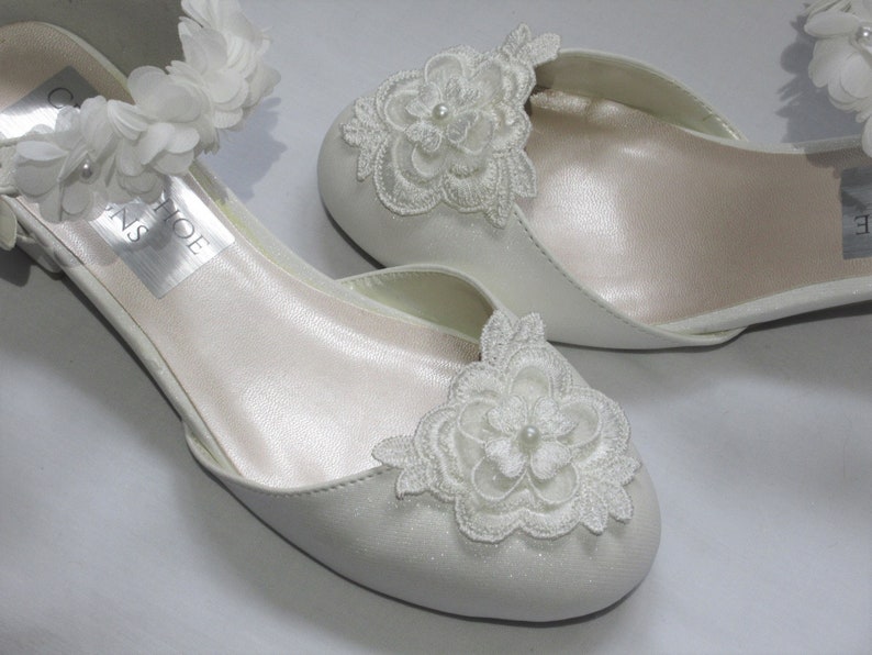 May include: White satin bridal shoes with delicate lace flower embellishments and an ankle strap adorned with white fabric flowers.