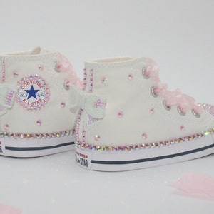 May include: White high-top Converse sneakers with pink bows and rhinestones. The sneakers have a white canvas upper, a black rubber sole, and a white star logo on the side. The words "Converse" and "All Star" are printed on the side of the shoe.