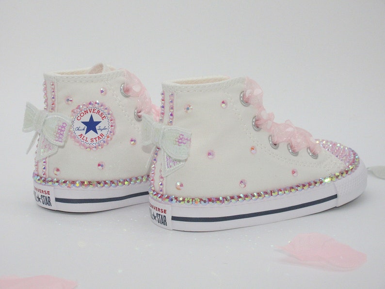 May include: White high-top Converse sneakers with pink bows and rhinestones. The sneakers have a white canvas upper, a black rubber sole, and a white star logo on the side. The words "Converse" and "All Star" are printed on the side of the shoe.