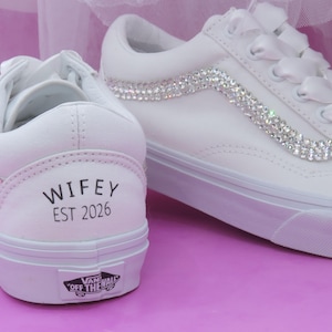 Könnte beinhalten: Weiße Sneaker mit Strassakzenten und Satinbandschnürsenkeln. Das Wort "WIFEY" und das Jahr "EST 2026" sind auf der Ferse eines Schuhs aufgedruckt. Die Schuhe haben eine weiße Sohle und das Vans-Logo.