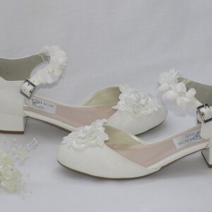 May include: A pair of white glitter heeled shoes with ankle straps and white flower embellishments. The shoes have a low block heel.