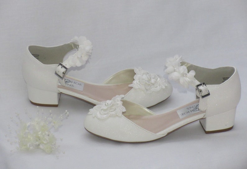 May include: A pair of white glitter heeled shoes with ankle straps and white flower embellishments. The shoes have a low block heel.