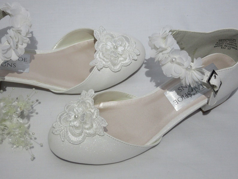 May include: White satin bridal shoes with a delicate floral design. The shoes have a buckle closure and a small heel. The brand name "Tailshoe Designs" is visible on the insole.