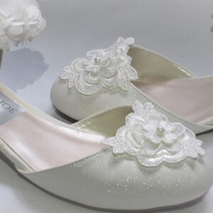 May include: A pair of white satin bridal shoes with a low heel. The shoes feature delicate white lace flower embellishments and a pearl accent. The shoes are labeled "Crystal Shoe Designs".