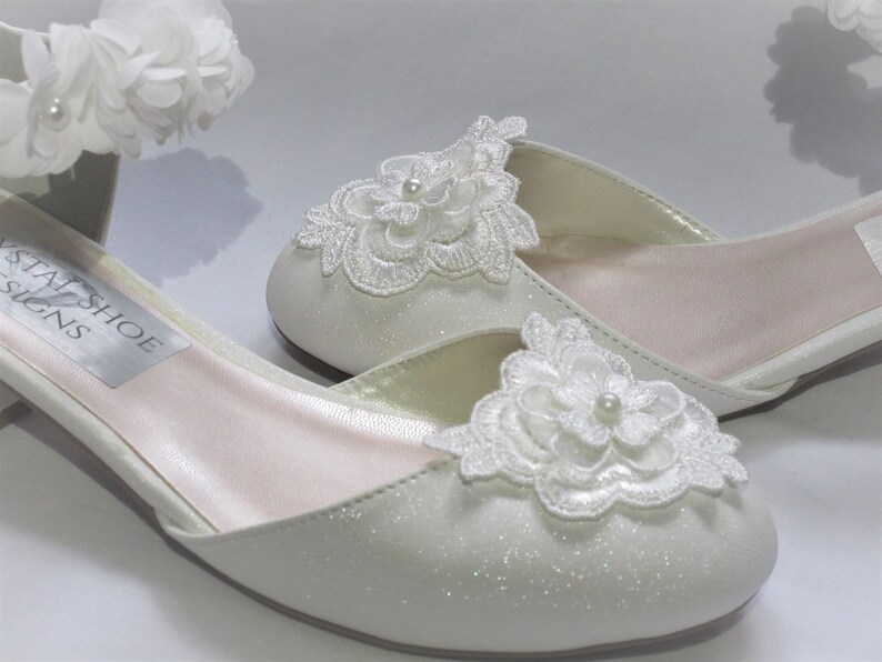 May include: A pair of white satin bridal shoes with a low heel. The shoes feature delicate white lace flower embellishments and a pearl accent. The shoes are labeled "Crystal Shoe Designs".