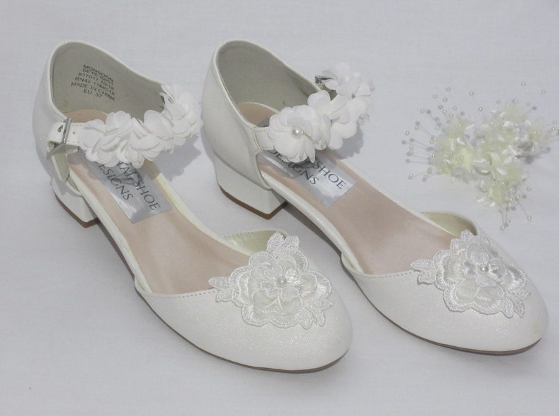 May include: White satin bridal shoes with a low heel and delicate lace flower embellishments. The shoes have a strap with a buckle closure and are adorned with white flowers.