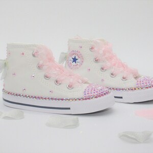 May include: White high-top sneakers with pink lace and rhinestones. The sneakers have a blue star logo on the side.