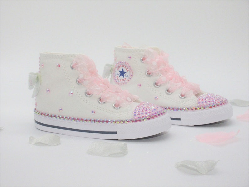May include: White high-top sneakers with pink lace and rhinestones. The sneakers have a blue star logo on the side.