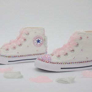 May include: White high-top Converse sneakers with pink lace and rhinestones. The sneakers have a blue star logo and the words "Converse All Star" on the side.