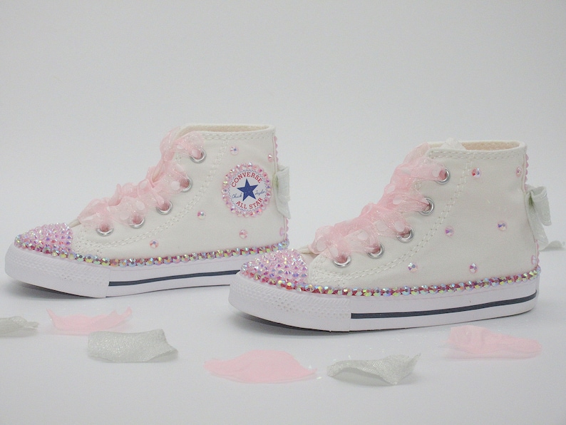 May include: White high-top Converse sneakers with pink lace and rhinestones. The sneakers have a blue star logo and the words "Converse All Star" on the side.