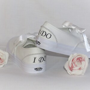 May include: White canvas trainers with satin ribbon laces and pearl detailing. The words "I DO" are printed in black on the heel of each shoe. Two pink roses are placed near the shoes.
