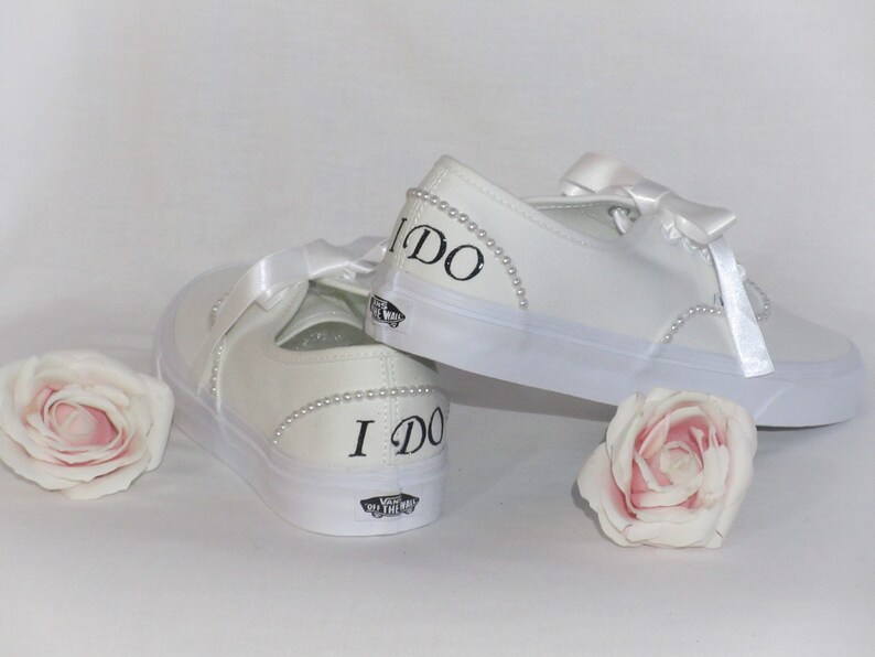 May include: White canvas trainers with satin ribbon laces and pearl detailing. The words "I DO" are printed in black on the heel of each shoe. Two pink roses are placed near the shoes.