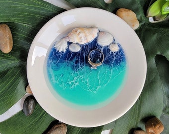 May include: White ceramic ring dish with a blue and turquoise resin ocean scene. The dish features seashells and a silver ring.
