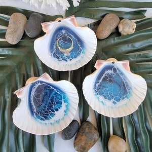 May include: Three white seashells with blue and white resin interiors. The shells are arranged on a green leaf background. One shell has a gold ring inside.