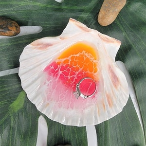 May include: A white seashell ring dish with a pink and orange resin design. A silver ring with a diamond is sitting in the dish.