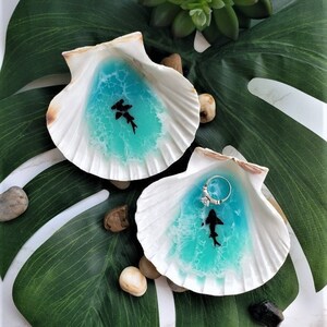 May include: Two white seashell ring dishes with blue resin and a black shark silhouette. The dishes are on a green leaf.
