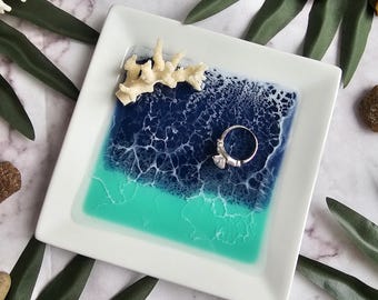 May include: Square white ceramic ring dish with a resin design in shades of blue and teal, resembling ocean waves. A silver ring with a clear stone rests on the resin. Decorative coral is affixed to the dish.