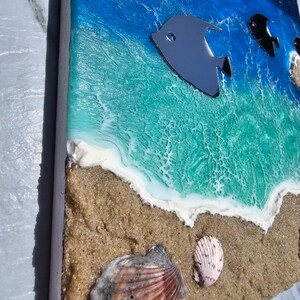 May include: Ocean-themed artwork featuring a beach scene with a blue and turquoise ocean, sandy shore, and seashells. Metallic fish accents swim in the water. The artwork is on a rectangular wooden panel.