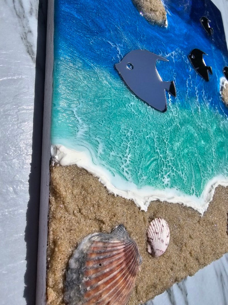 May include: Ocean-themed artwork featuring a beach scene with a blue and turquoise ocean, sandy shore, and seashells. Metallic fish accents swim in the water. The artwork is on a rectangular wooden panel.