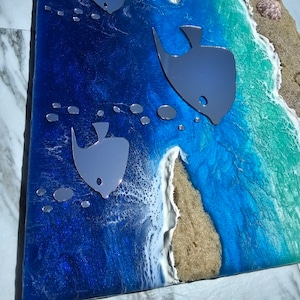 Puede incluir: Una pieza de arte de resina azul y verde con una escena de playa. La pieza presenta olas blancas, arena marr&oacute;n y tres peces plateados. Los peces nadan en el agua azul.