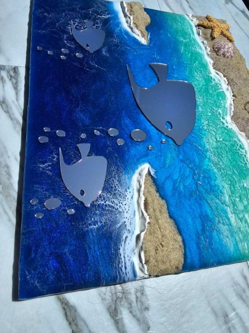 Puede incluir: Una pieza de arte de resina azul y verde con una escena de playa. La pieza presenta olas blancas, arena marr&oacute;n y tres peces plateados. Los peces nadan en el agua azul.
