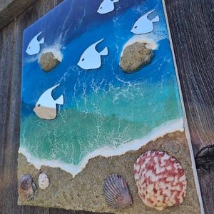 May include: A square, coastal-themed artwork featuring a vibrant ocean scene. White fish swim above a sandy beach with seashells. The ocean transitions from deep blue to turquoise, with foamy white waves.