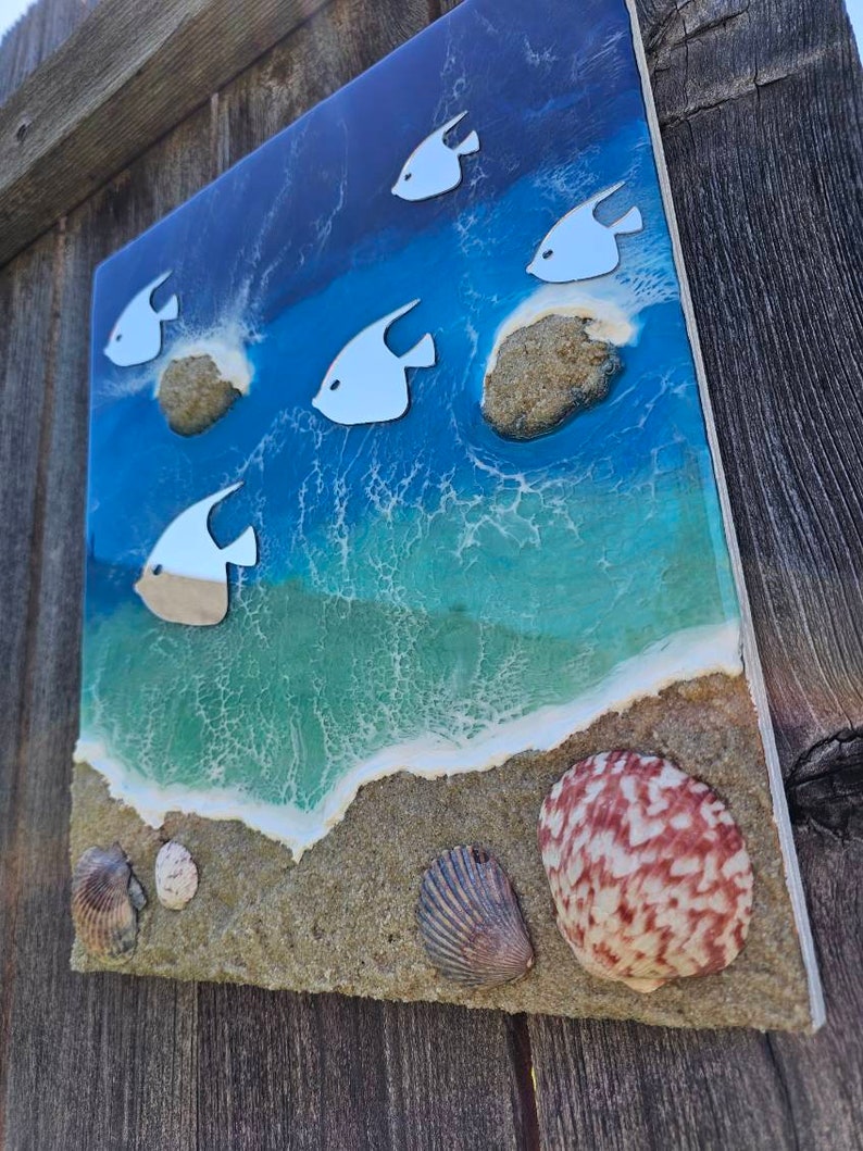 May include: A square, coastal-themed artwork featuring a vibrant ocean scene. White fish swim above a sandy beach with seashells. The ocean transitions from deep blue to turquoise, with foamy white waves.
