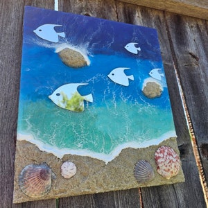 May include: A rectangular beach-themed artwork featuring a vibrant ocean scene. White fish swim above the waves. The artwork incorporates sand, seashells, and a textured surface, creating a three-dimensional effect. The color palette includes shades of blue, green, and tan.