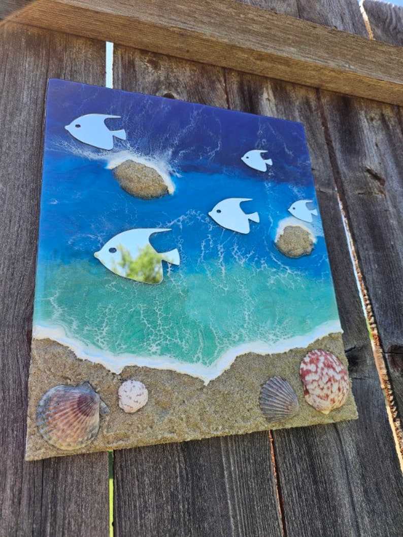 May include: A rectangular beach-themed artwork featuring a vibrant ocean scene. White fish swim above the waves. The artwork incorporates sand, seashells, and a textured surface, creating a three-dimensional effect. The color palette includes shades of blue, green, and tan.