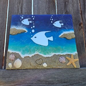 Puede incluir: Una pieza de arte de resina azul y blanca que representa una escena de playa con peces blancos, conchas y una estrella de mar en una playa de arena.