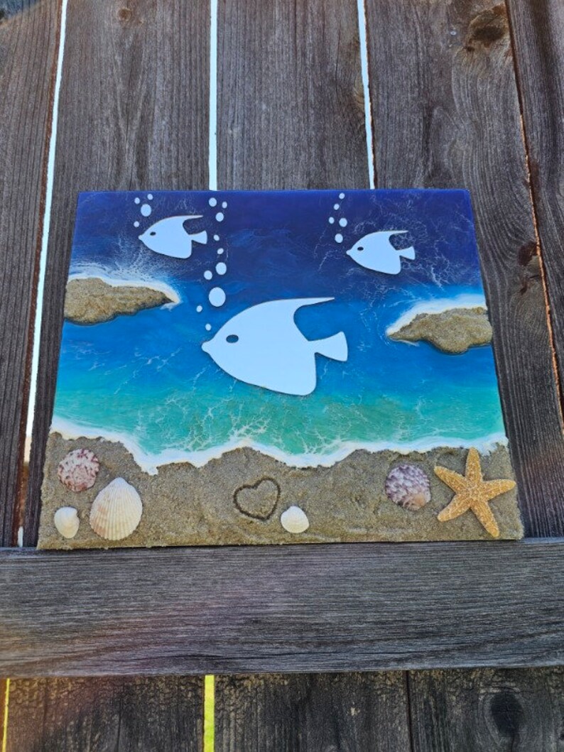 Puede incluir: Una pieza de arte de resina azul y blanca que representa una escena de playa con peces blancos, conchas y una estrella de mar en una playa de arena.