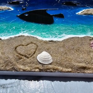 Puede incluir: Primer plano de una escena de playa con un coraz&oacute;n dibujado en la arena, una concha blanca y una concha rosa. El fondo es un oc&eacute;ano azul con una ola turquesa y dos peces negros.