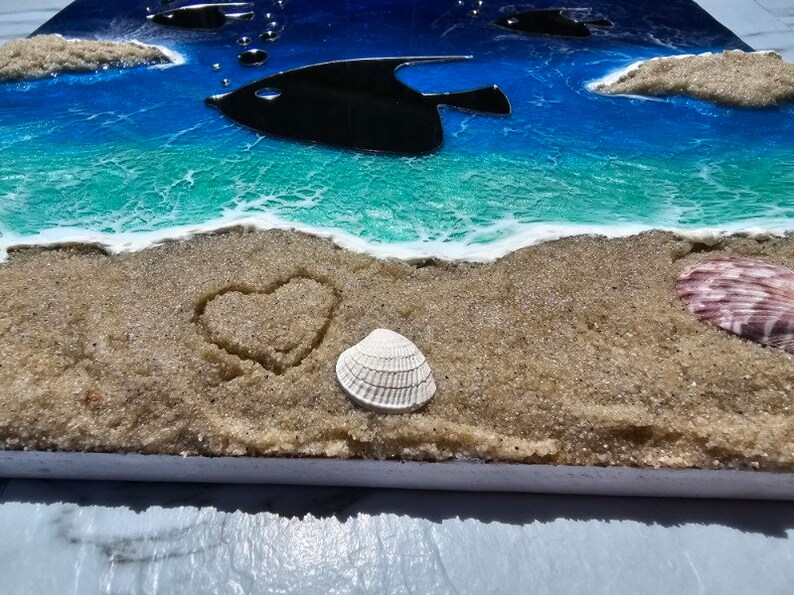 Puede incluir: Primer plano de una escena de playa con un coraz&oacute;n dibujado en la arena, una concha blanca y una concha rosa. El fondo es un oc&eacute;ano azul con una ola turquesa y dos peces negros.
