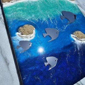 May include: A rectangular resin art piece depicting a beach scene. The artwork features a sandy shore, turquoise waves, and deep blue ocean with silver fish accents. Seashells are embedded in the sand.